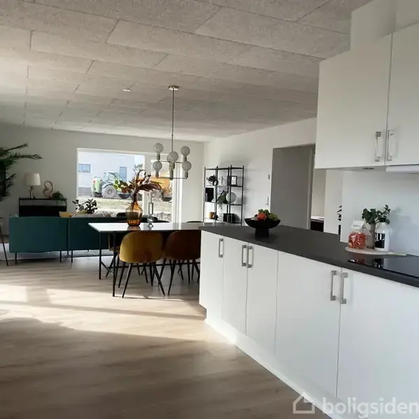 Køkken med hvide skabe og sort bordplade fører til en spisestue med farverige stole omkring et bord. Store vinduer og moderne indretning præger rummet.