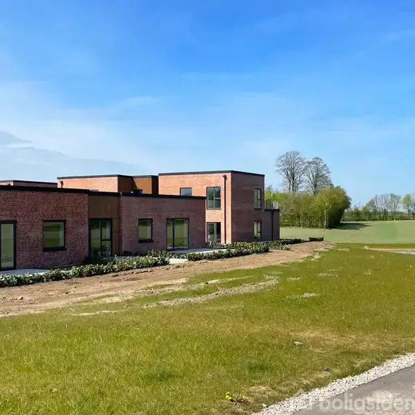 En moderne, lav, murstensbygning står stille i en grøn mark omkranset af lave buske. I baggrunden ses en blå himmel og træer ved horisonten.