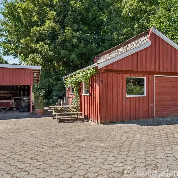 Rødt træskur med garageport står ved siden af et halvtag, hvor en trailer er parkeret. Omkranset af grønne træer og buske, foran bygningen står et træbord med bænk.