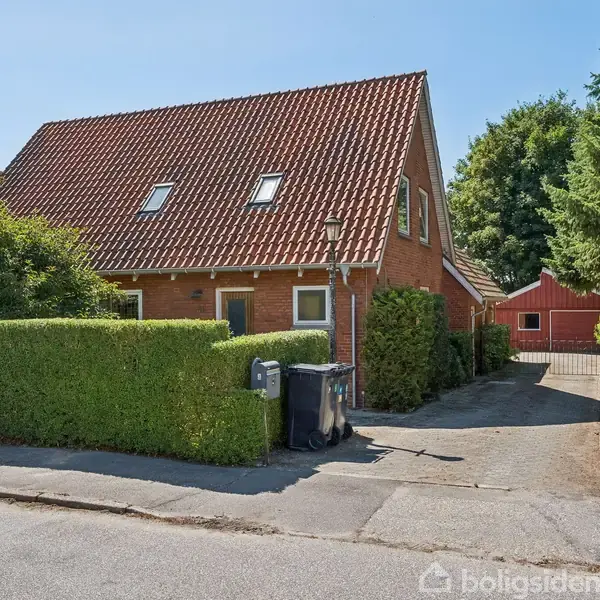 Hus med rødt tegltag og to skraldespande, står ved en hæk i en villavej. Der er en indkørsel til venstre og træer rundt omkring.