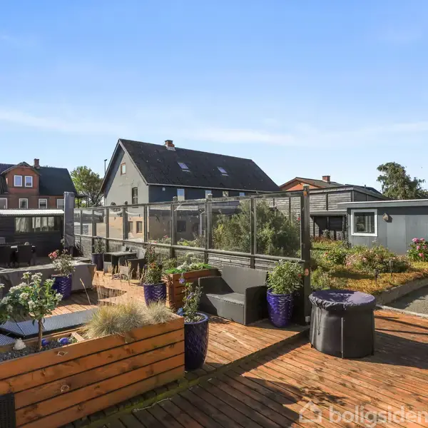 Træterrasse omringet af blomsterkasser og træmøbler. Terrassen befinder sig i en baggård med udsigt til omkringliggende huse og en klar, blå himmel.