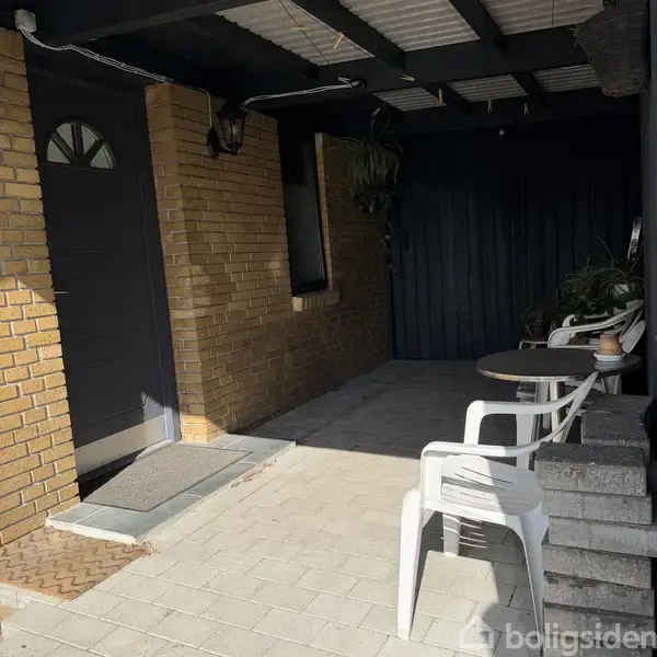 En dør omgivet af murstensvægge fører ind til et hus. To hvide plasticstole står ved et rundt bord på en flisebelagt terrasse ved siden af blomsterkrukker.