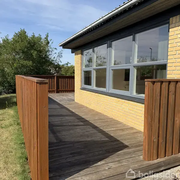 Træterrasse med gelænder omgiver en gul murstensbygning med store vinduer. Grønt græs og træer ses i baggrunden under en blå himmel.