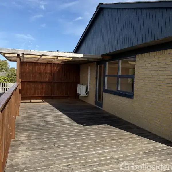 Terrasse med trædæk og trægelænder, dækket af et halvtag. Murstensbygning med stort vindue til højre. Solskin og klar blå himmel i baggrunden.