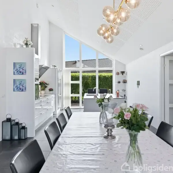 Langt spisebord med en dug og blomsterdekorationer står i et lyst og moderne køkken-alrum. Store vinduer giver udsigt til en have. En elegant lysekrone hænger over bordet.
