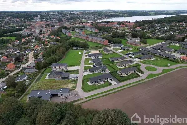 Dronebillede af en moderne boligudvikling; huse med flade tage og grønne græsplæner er arrangeret langs veje. I baggrunden ses en by med røde bygninger og en sø.