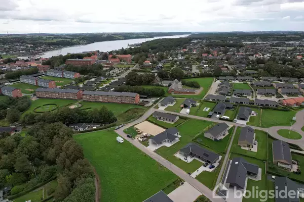 Luftfoto af et boligområde med huse omgivet af grønne marker. En flod strækker sig gennem landskabet i baggrunden, og mange træer er spredt i området.