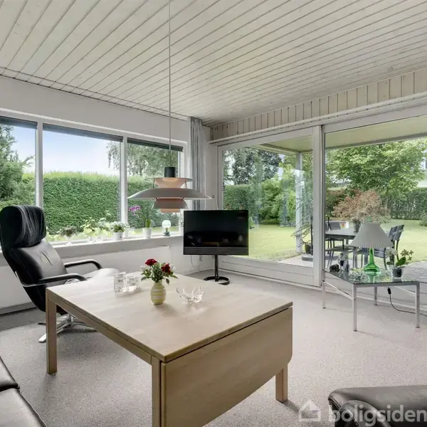 Stuen har en sort lædersofa og en lænestol, omgivet af hvide vægge og store vinduer med udsigt til en frodig have. En glasdør fremhæver rummets lyse atmosfære.