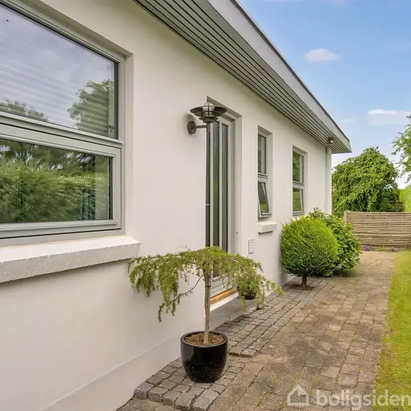 Et hvidt hus med store vinduer står langs en stenbelagt gangsti. Foran huset er der plantekrukker og en klippet busk. Græsplæne og grøn hæk omkranser området.