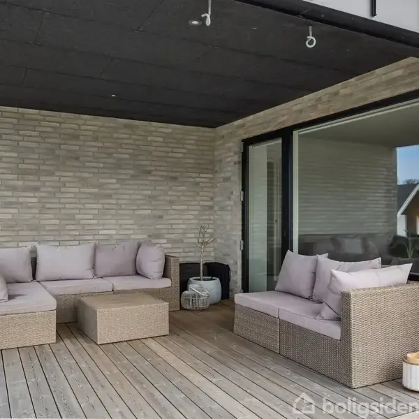 En udendørs terrasse med lysebrune fletsofaer med puder står på et trædæk. En glasdør fører til indendørs. To lanterner er placeret i hjørnet.