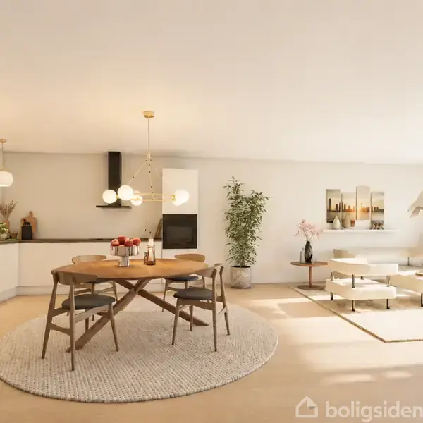 Rundt spisebord med stole står på tæppe, omgivet af moderne køkken og stue med lys indretning, sofaer, planter og dekorative elementer. Boligen virker lys og rummelig med enkelt design.