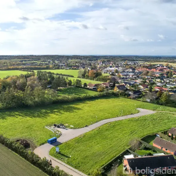 Grønt græsareal omgivet af en snoet vej; en blå container står på vejen. I baggrunden ses en række huse og en by mod horisonten under en overskyet himmel.