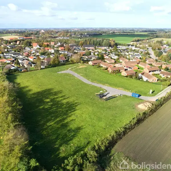 En droneoptagelse viser en grøn mark med en asfaltvej, der snor sig gennem landskabet. I baggrunden ses en by med huse omgivet af marker og træer.
