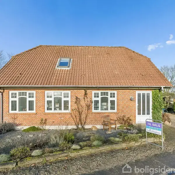 Et rødt murstenshus med rødt tegltag står stille i en have med buske og grussti. Et skilt ved fortovet siger "TIL SALG", indikerende en ejendom til salg.