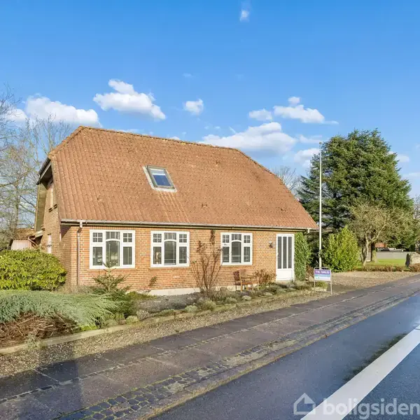 Et rødt murstenshus med skråt tegltag står ved siden af en vej. Haven er omkranset af buske. I baggrunden er der træer og en klar blå himmel med få skyer.