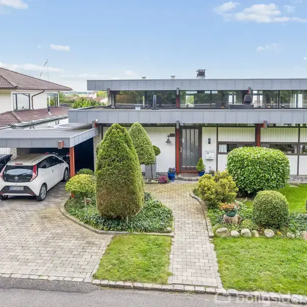 En moderne bolig med fladt tag, hvid bil parkeret i carporten. Foran huset er der en velplejet have med klippede buske. Det er en solrig dag med få skyer.