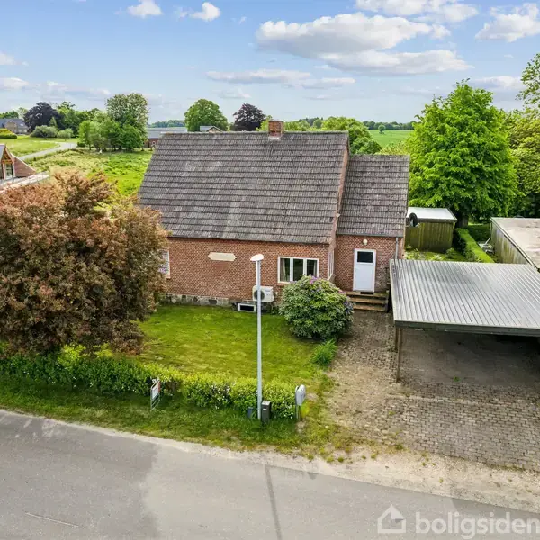 Murstenshus med gråt tag, der står stille omgivet af grønne træer og buske. Det har en indkørsel med carport og ligger ud til en vej i et landligt område.