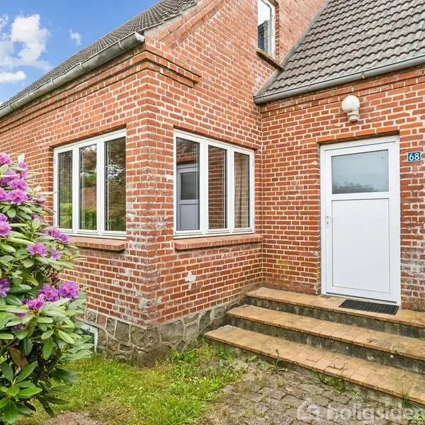 Rødt murstenshus med hvide vinduer står stille ved siden af blomstrende lilla busk. Indgangen består af en hvid dør med trappe foran. Solrigt vejr og blå himmel. Husnummer skilt: 48.