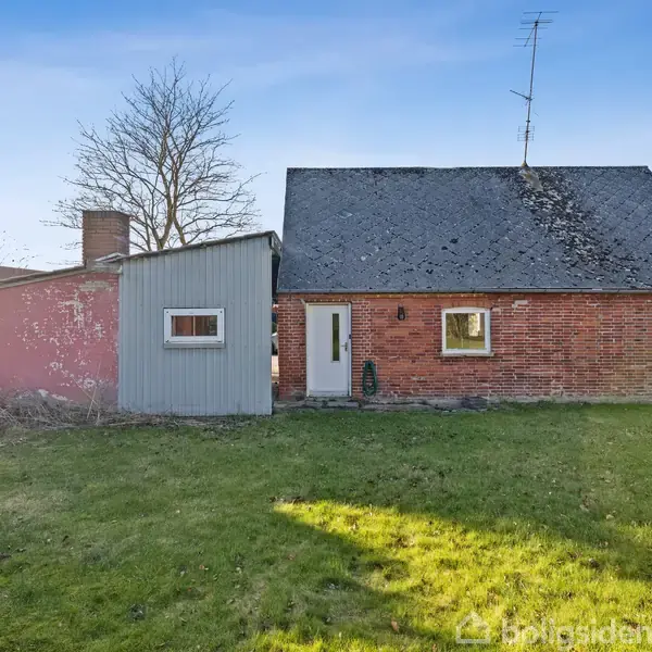Et murstenshus med skråt tag står på en græsplæne. Ved siden af huset er et lille skur. Himlen er blå, og området er grønligt og åbent.