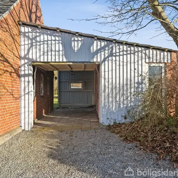 Bygning med rød murstens- og hvid pladefacade har en åben passage. Omgivet af grusbelagt jord og et træ til højre. Fliser danner gulvet under passagen.