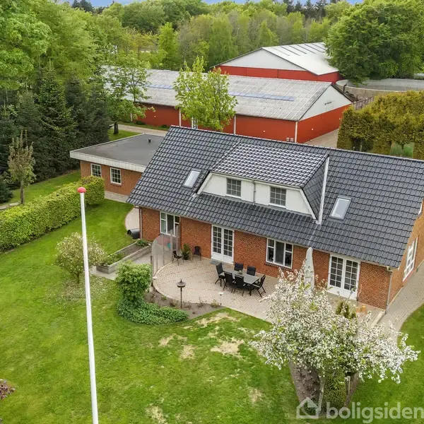 En rødstensvilla med gråt tegltag står omgivet af grønne træer og en have. En terrasse med havemøbler ses foran huset, mens et stort rødt udhus er placeret i baggrunden.