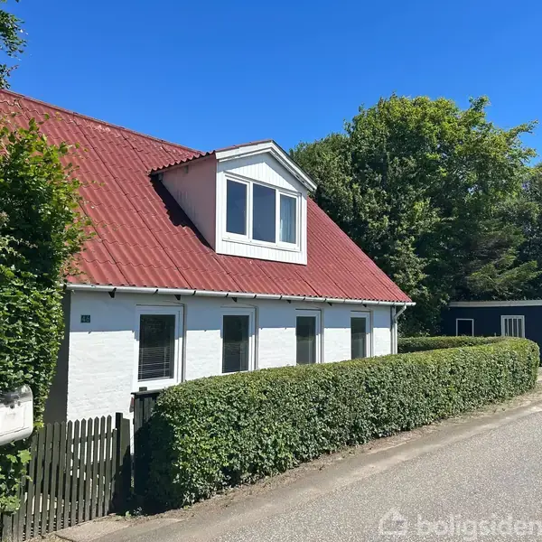 Et hvidt hus med rødt tag står langs en vej. Buske omgiver grunden. Blå himmel og træer skaber en sommerlig atmosfære.
