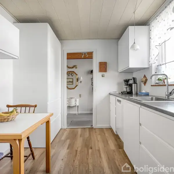Et gult murstenshus med en flad tagkonstruktion står stille i en grøn have omgivet af hække. En indkørsel leder op til en garage på husets venstre side.