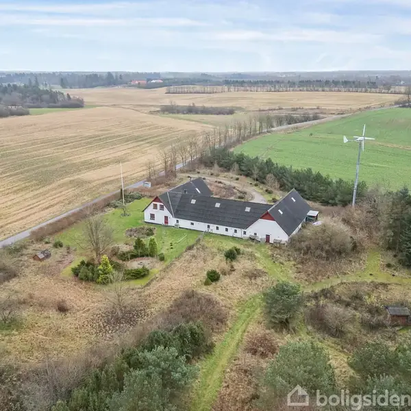 Et hvidt landhus med sort tag står omgivet af marker og grønne områder. En vindmølle ses ved siden af huset, og en lang landevej strækker sig langs ejendommen.