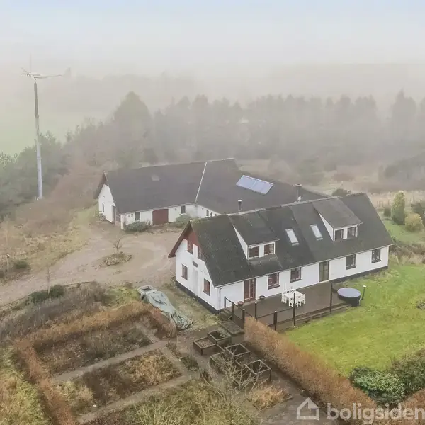 Et landhus med solpanel på taget ligger i en landlig have. Omkring huset er der stier, buske og et skur. Baggrunden er præget af træer og en vindmølle i tåge.