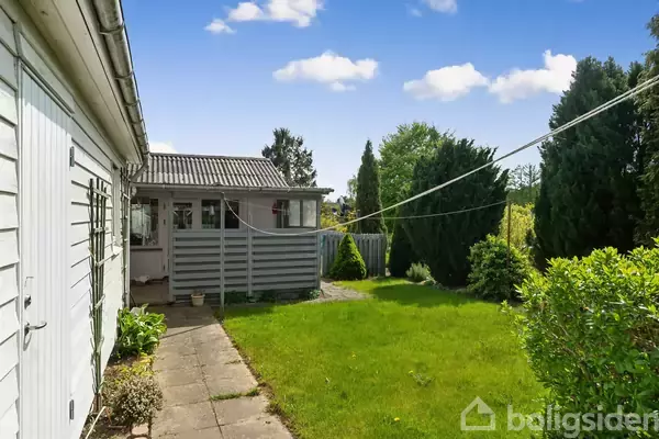 Et hus med hvidt træpanel står ved siden af en grøn græsplæne med buske. En flisebelagt sti leder fra terrassen gennem haven under en blå himmel med få skyer.