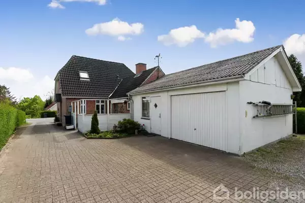 Et hus med en stor garage ligger i en stille boligområde. Huset har en rød murstensfacade, sort tegltag, og en indkørsel med fliser omgivet af hæk og haveplanter.