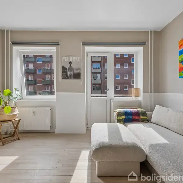 Stue med sofa, tv på væggen og maleri; lysindfald fra to vinduer og altandør. Plakat med teksten "PIAZZOLLA" over en radiator ved siden af en grøn plante på et lille bord.