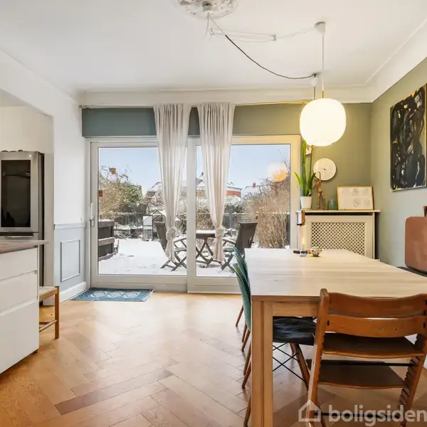 Et køkken-alrum med spisebord og stole står foran en stor skydedør med udsigt til en terrasse. Væggene har kunstværker, og der hænger en rund lampe ovenover.