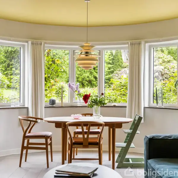 Rundt spisebord med stole står foran en række buede vinduer med udsigt til frodig have. Over bordet hænger en elegant lampe. Planter pryder vindueskarmen.
