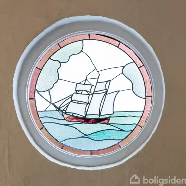 Rundt farvet glasvindue viser et sejlskib på havet med skyer i baggrunden, sat i en hvid ramme på en beige væg.