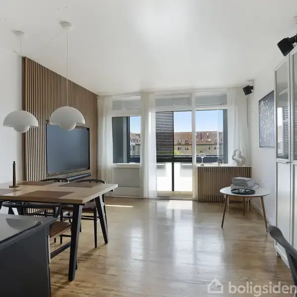 Spisestue med bord og stole, fladt TV på væggen. Store vinduer lader lys ind, moderne design med trægulv og hvide skabe. Pæne pendellamper hænger over bordet.