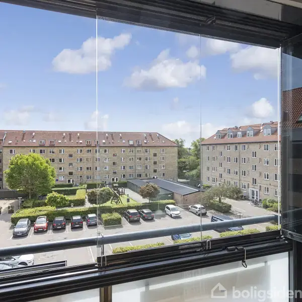 Fra en glasinddækket balkon ses en udsigt over en gård med parkerede biler og to boligblokke med røde tage. Himlen er klar med spredte skyer.