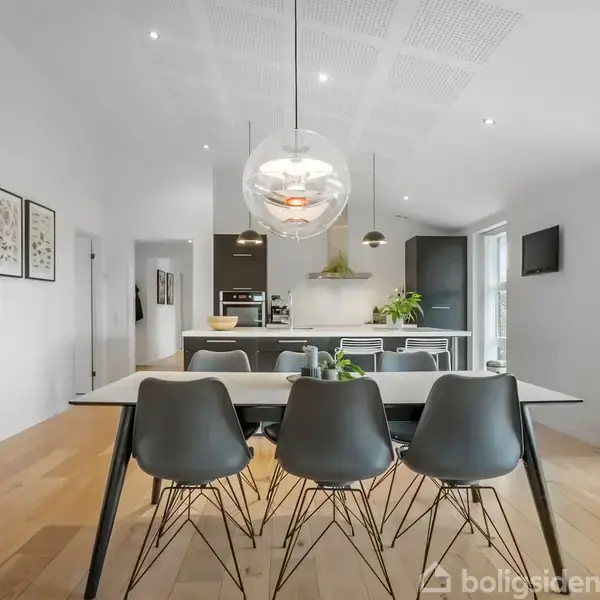 Spisebord med fire stole står i et åbent køkken-alrum. Over bordet hænger en stor, rund lampe. Væggene er lyse med indrammede billeder, og der er store vinduer til højre.