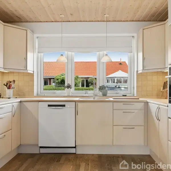 Køkken med lyse skabe og træloft. Opvaskemaskine og vask er centralt placeret ved vindue. Udenfor ses et hus med rødt tag. Indretning er lys og modern.