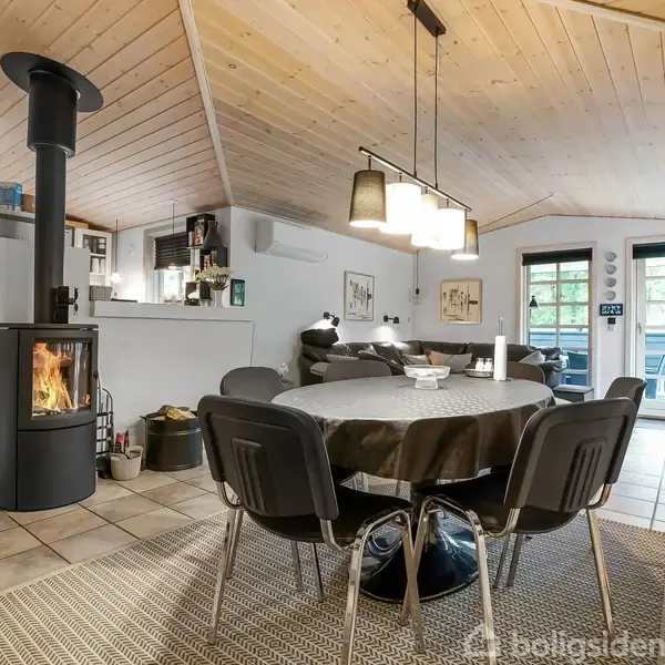Et rundt spisebord dækket med en mørk dug står midt i en stue med åben tagkonstruktion. En brændeovn brænder ved siden af, og der er en sofa i baggrunden.