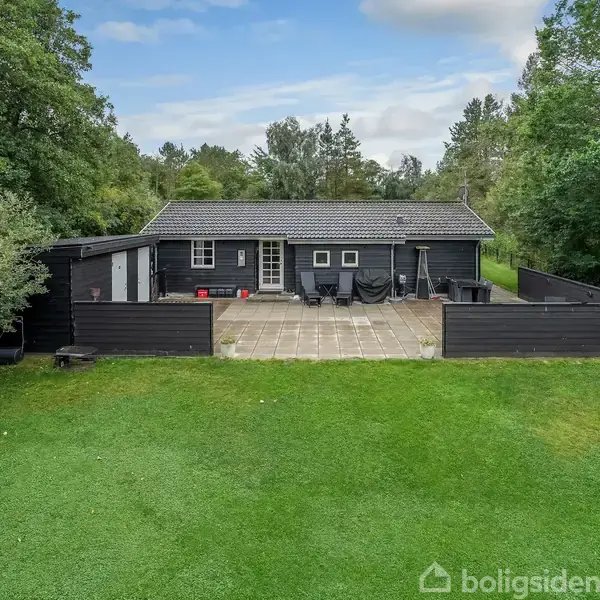 Et sort træhus står stille, omgivet af grønne træer og en græsplæne. Foran huset er der en stor, stenbelagt terrasse med havemøbler.
