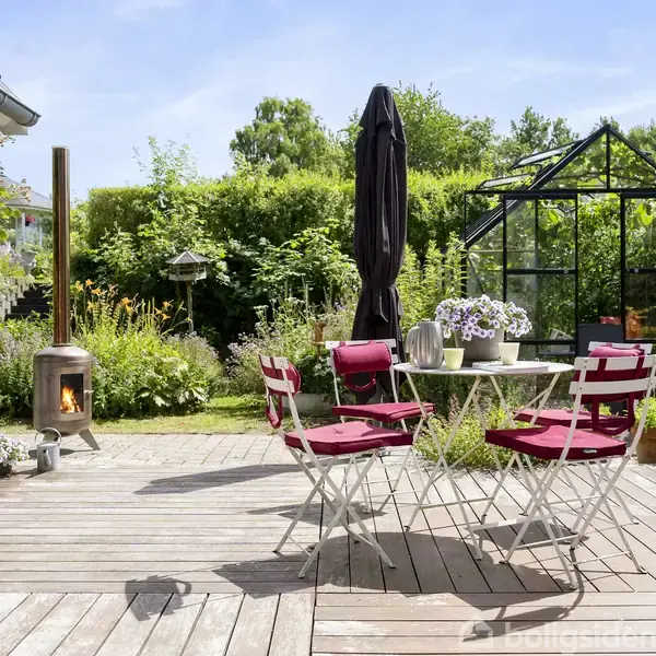 Bord med røde stole står på en træterrasse med blomsterdekorationer. Ved siden af ses en brændeovn ved siden af buske og en havepavillon. Solrig dag i en frodig have.