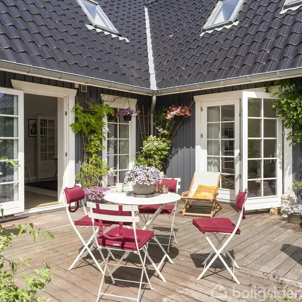 Terrassemøbler med røde hynder står på en træterrasse foran et hus med mørk tag og hvide døre. Planter og blomster pynter terrassen, der bades i sollys.