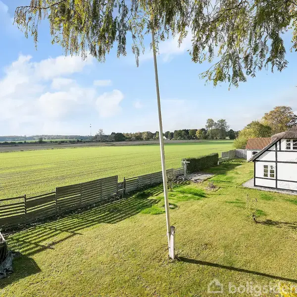 Et bindingsværkshus står stille i en frodig have, omgivet af åbne marker under en klar blå himmel, med en flagstang og hegn langs græsplænen.