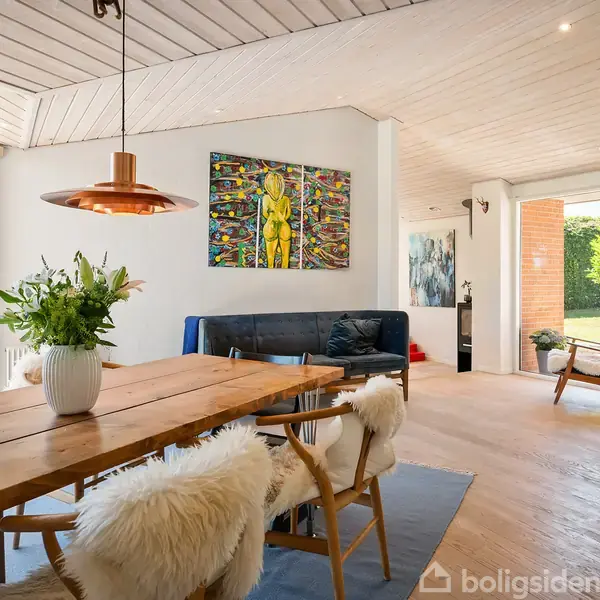 Langt træbord med blomsterbuket står i spisestue; stole med hvidt stof omkring. Kobberlampe hænger over. Bagved ses en mørk sofa, kunst på væggen, og stor dør til have.