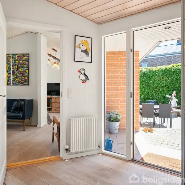 Indgangsparti med åben dør mod terrasse. Fuglebilleder pryder væggen. Indendørs ses radiator og åbning til stue med sofa. Udendørs terrasse har møbler og grøn hæk.