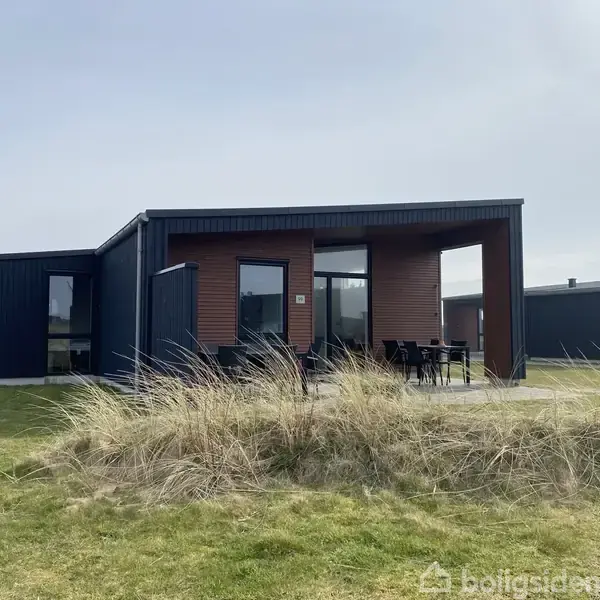 Et moderne hus står i et græsbeklædt område, med store vinduer og terrasse med møbler. Bygningen har en flad taglinje og enkle, geometriske former.