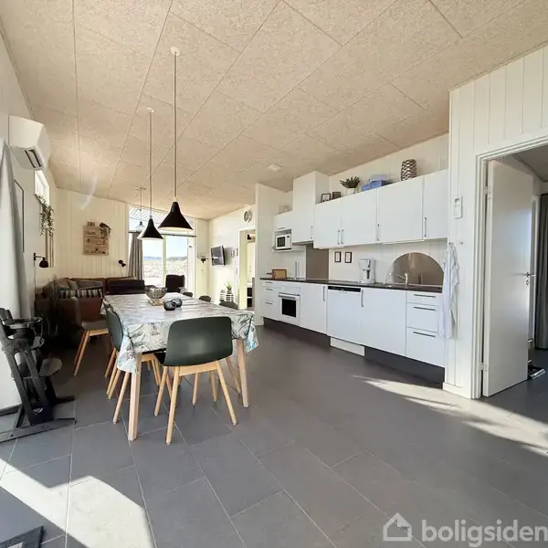 Spisebord med stole står i lys stue-køkken-alrum. Rummet har store vinduer, to lamper over bordet, moderne køkkenudstyr på en lang hvid bordplade, og en sofa i baggrunden.