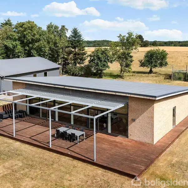 En moderne bungalow med fladt tag står på en stor træterrasse. Ved siden af ses en garage. Omgivelserne består af en græsmark med træer i baggrunden.