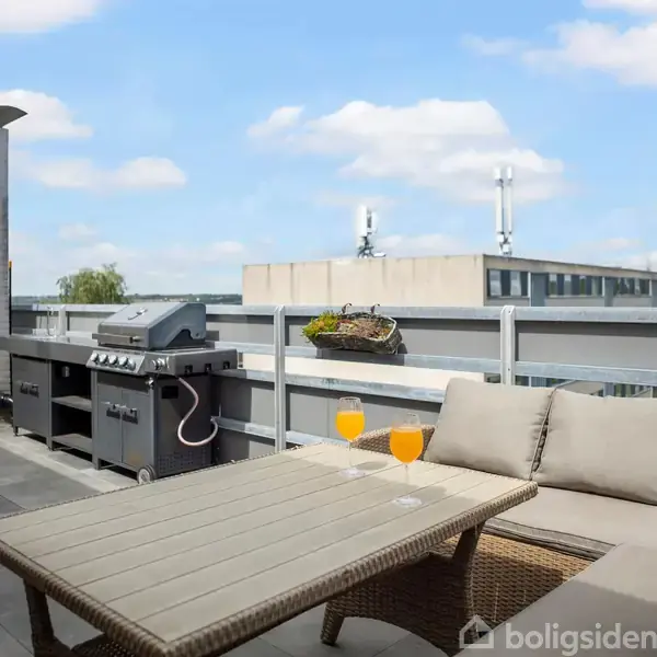 Grill står ubrugt på terrasse ved siden af moderne sofa og bord med to glas orange drikke. Omgivet af byudsigt og blå himmel med få skyer.